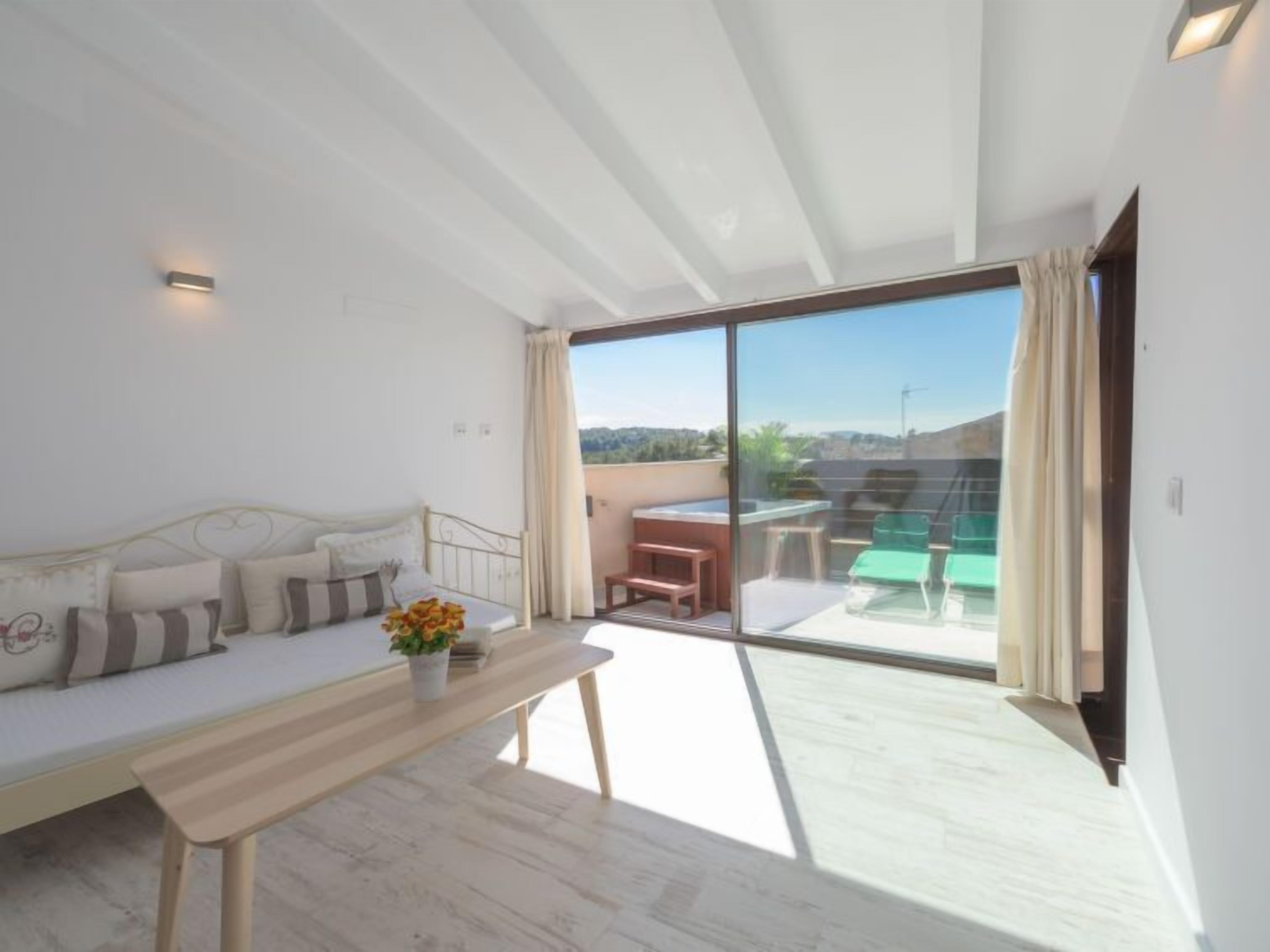 A modern bedroom with a sofa, a bedside table with a vase of flowers, and a large sliding glass door leading to a balcony with outdoor furniture.