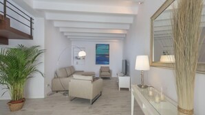 Uma sala de estar moderna com um sofá, uma mesa de centro, uma televisão e um abajur. Há uma escada à esquerda e um espelho grande à direita.