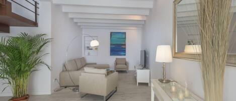 A modern living room with a sofa, a coffee table, a TV, and a lamp. There is a staircase on the left and a large mirror on the right.