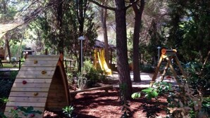 A playground with a wooden slide, climbing wall, and swings set in a wooded area.