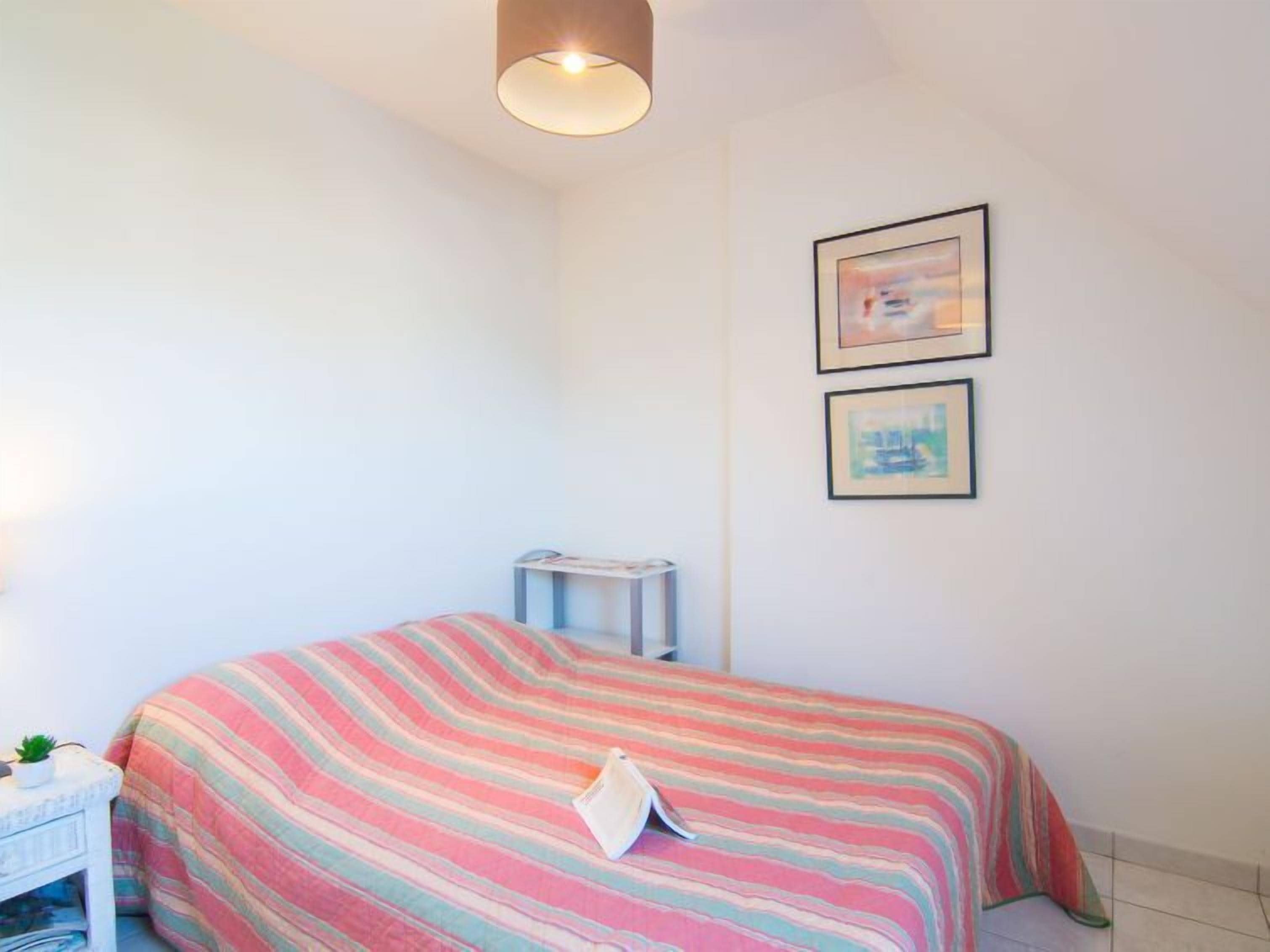 A bedroom with a bed, a small bedside table with a plant, and two framed pictures on the wall.