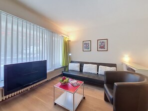 A modern living room with a flat-screen TV, a sofa with cushions, a coffee table with fruits, and a chair.