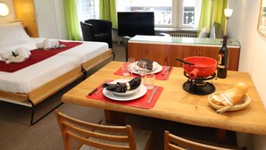 Hotellihuone, jossa on sänky, ruokapöytä, jonka päällä on lautasia, laseja ja viinipullo. Taustalla näkyy tuoli, työpöytä ja toinen tuoli.