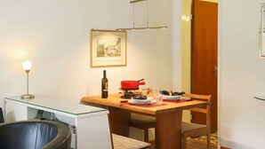 Hotellihuone, jossa on kahdelle katettu ruokapöytä, tuoli, lamppuvarustettu työpöytä, seinällä kehystetty kuva ja ovi, joka johtaa toiseen huoneeseen.
