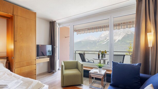 Ein Hotelzimmer mit Bett, Sofa, Fernseher, einem Balkon mit Tisch und Stühlen sowie Blick auf die Berge.