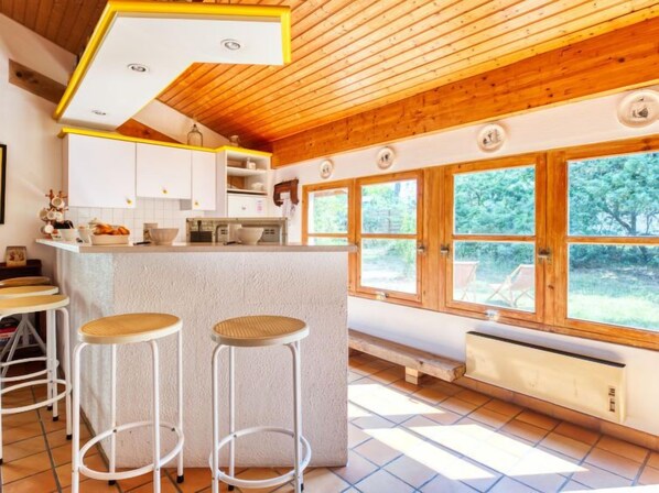 A kitchen with wooden ceiling, white cabinets, and a large window. There are two stools and a radiator.