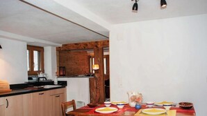 Ein Essbereich mit einem Holztisch, gedeckt für vier Personen, umgeben von Holzsesseln. Im Hintergrund ist eine Küche mit Holzausbau und einem Fenster erkennbar.