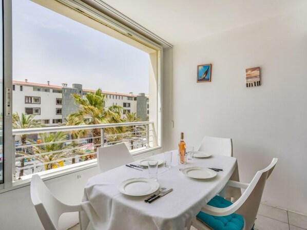 A dining area with a table set for two, a window with a view of buildings and palm trees, and two small paintings on the wall. - Le Sunset/Cap Sud by Interhome (Cap d'Agde)