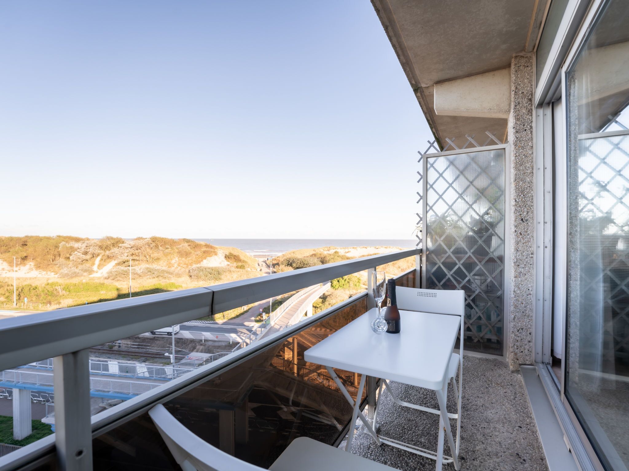 A balcony with a white table and chair, overlooking a coastal landscape with hills and a road.