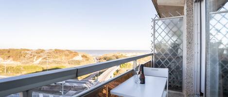 A balcony with a white table and chair, overlooking a coastal landscape with hills and a road.