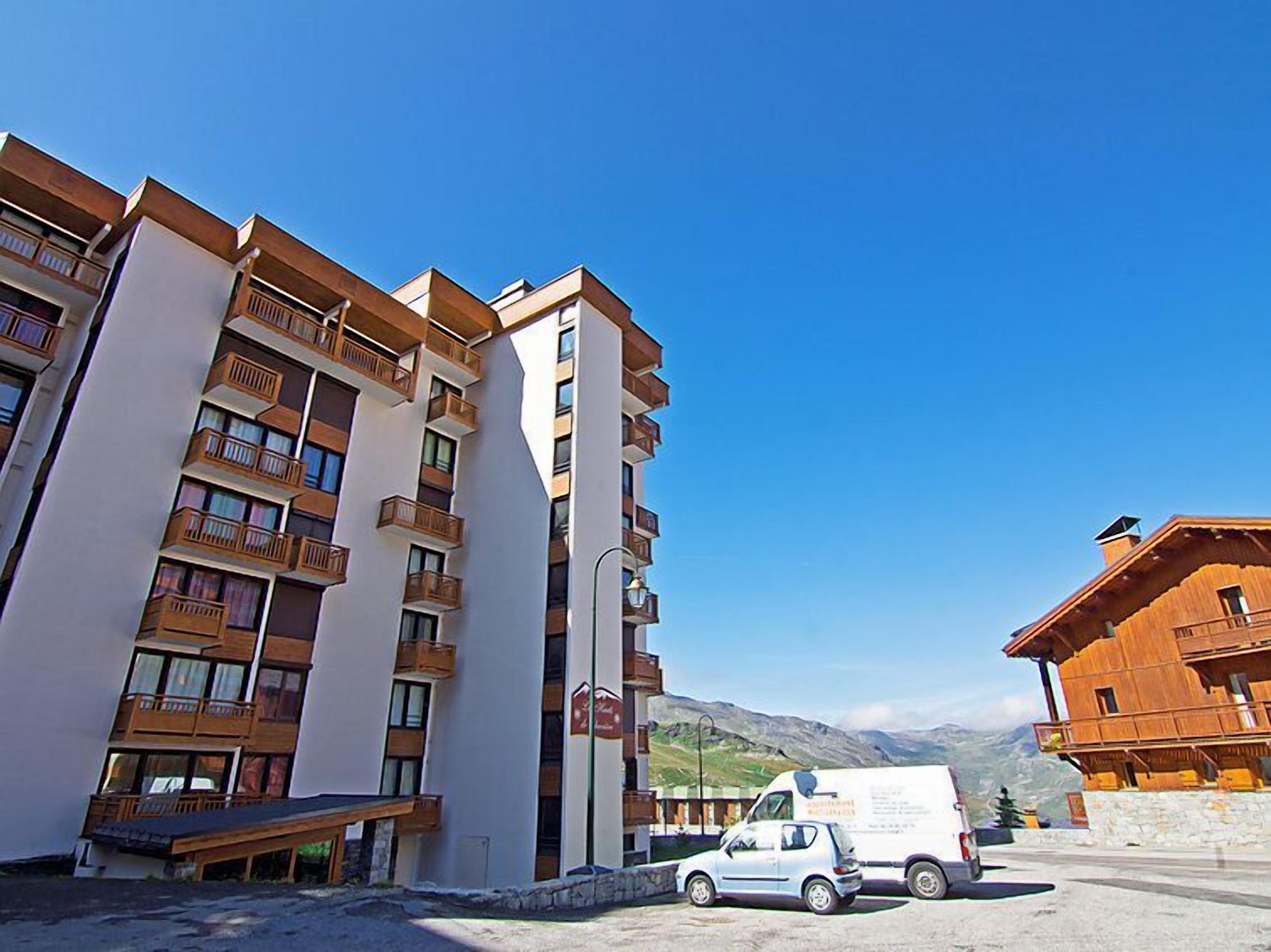 A multi-story hotel with balconies, a parking area, and a mountainous backdrop.