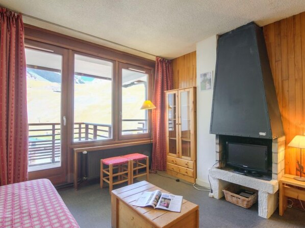 A cozy room with a bed, a wooden table with a magazine, a TV on a stone fireplace, and large windows with curtains. - Le Curling B (Val Claret) by Interhome (Tignes)