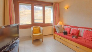 A room with a television, a wicker chair with a yellow cushion, a red sofa with patterned pillows, and a window with curtains. - Le Curling B (Val Claret) by Interhome (Tignes)