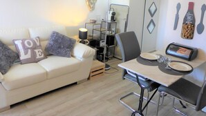 A modern living room with a white sofa, a dining table with chairs, a coffee machine, and a wall clock. - Kennedy by Interhome (Villers sur mer)