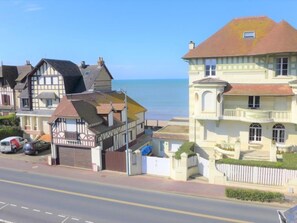 A coastal street with traditional European architecture, including a two-story building with a balcony and a corner house with a sloped roof.