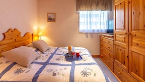 Une chambre avec un plafond en bois, un lit recouvert d’une courtepointe à motifs floraux, une table de chevet avec une lampe, une fenêtre avec des rideaux et des armoires en bois.