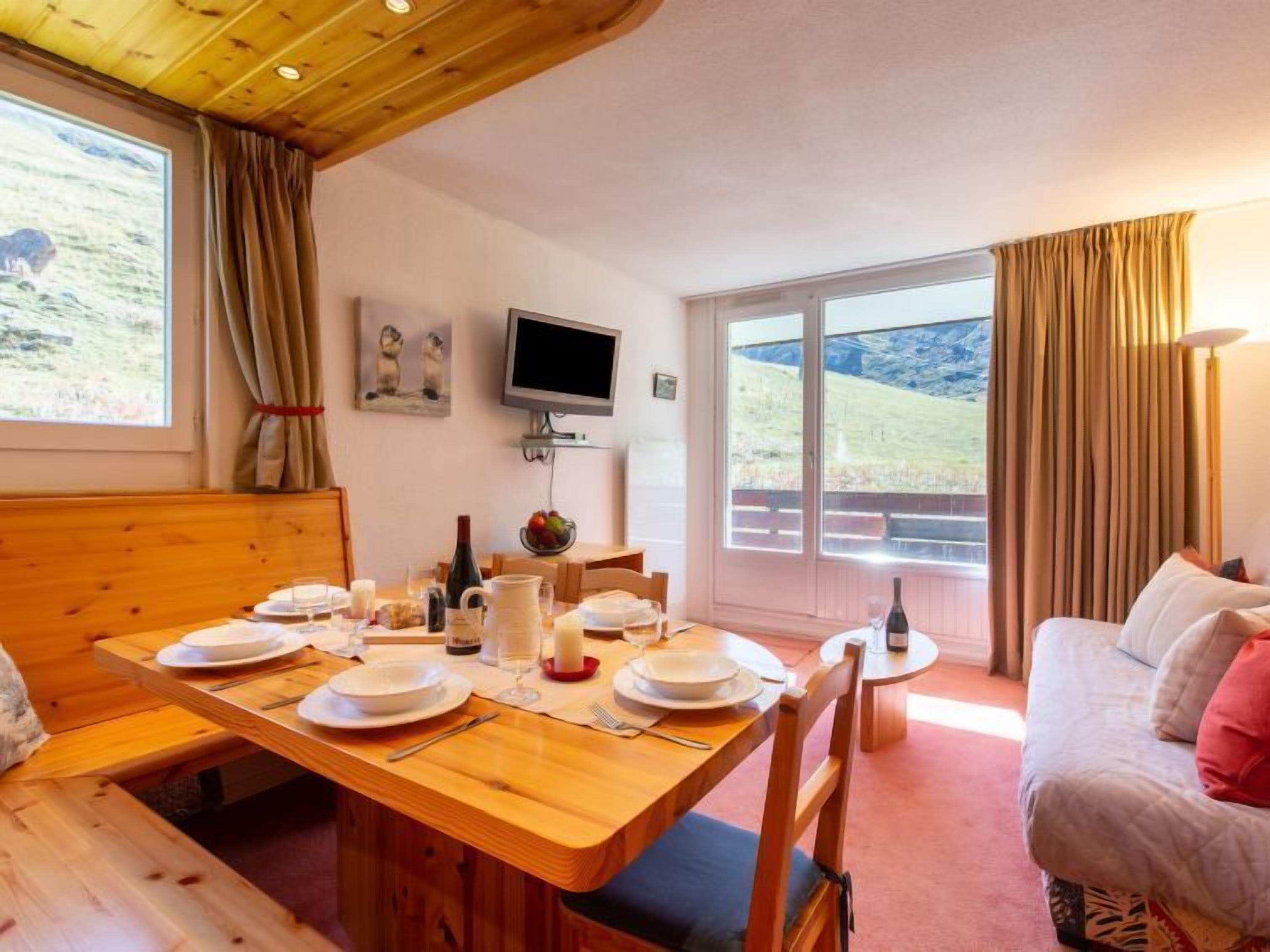 A cozy dining area with a wooden table set for four, a television on the wall, and a scenic view through the large windows.