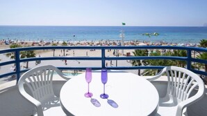Un balcon avec une table blanche et deux verres à vin violets, donnant sur une plage où se trouvent des gens et où flotte un drapeau.