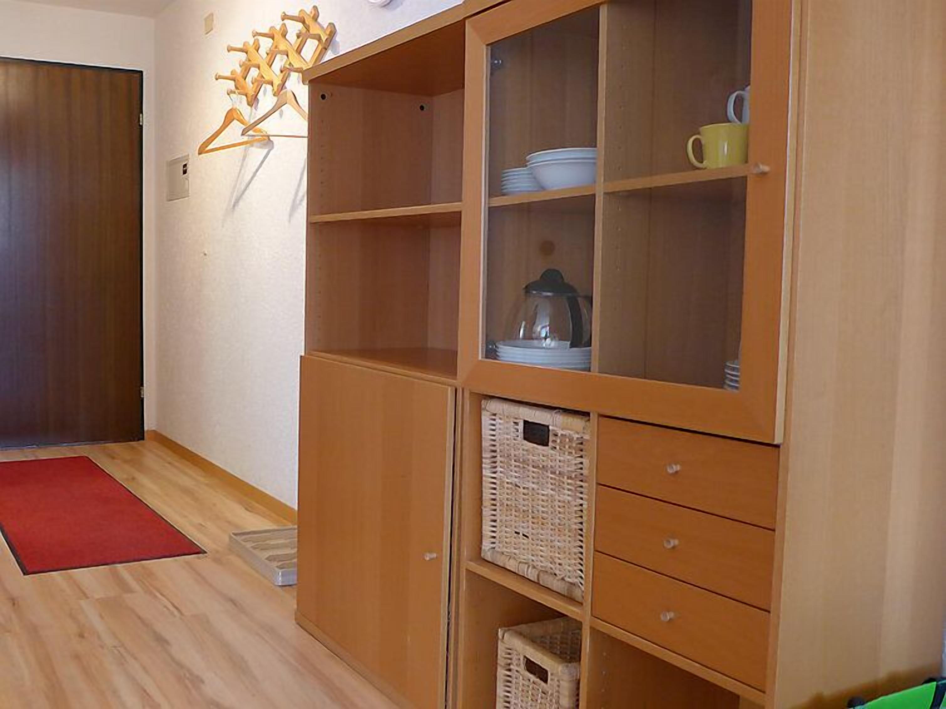 A room with a wooden cabinet containing dishes and a yellow mug, a red rug, and a wooden door.