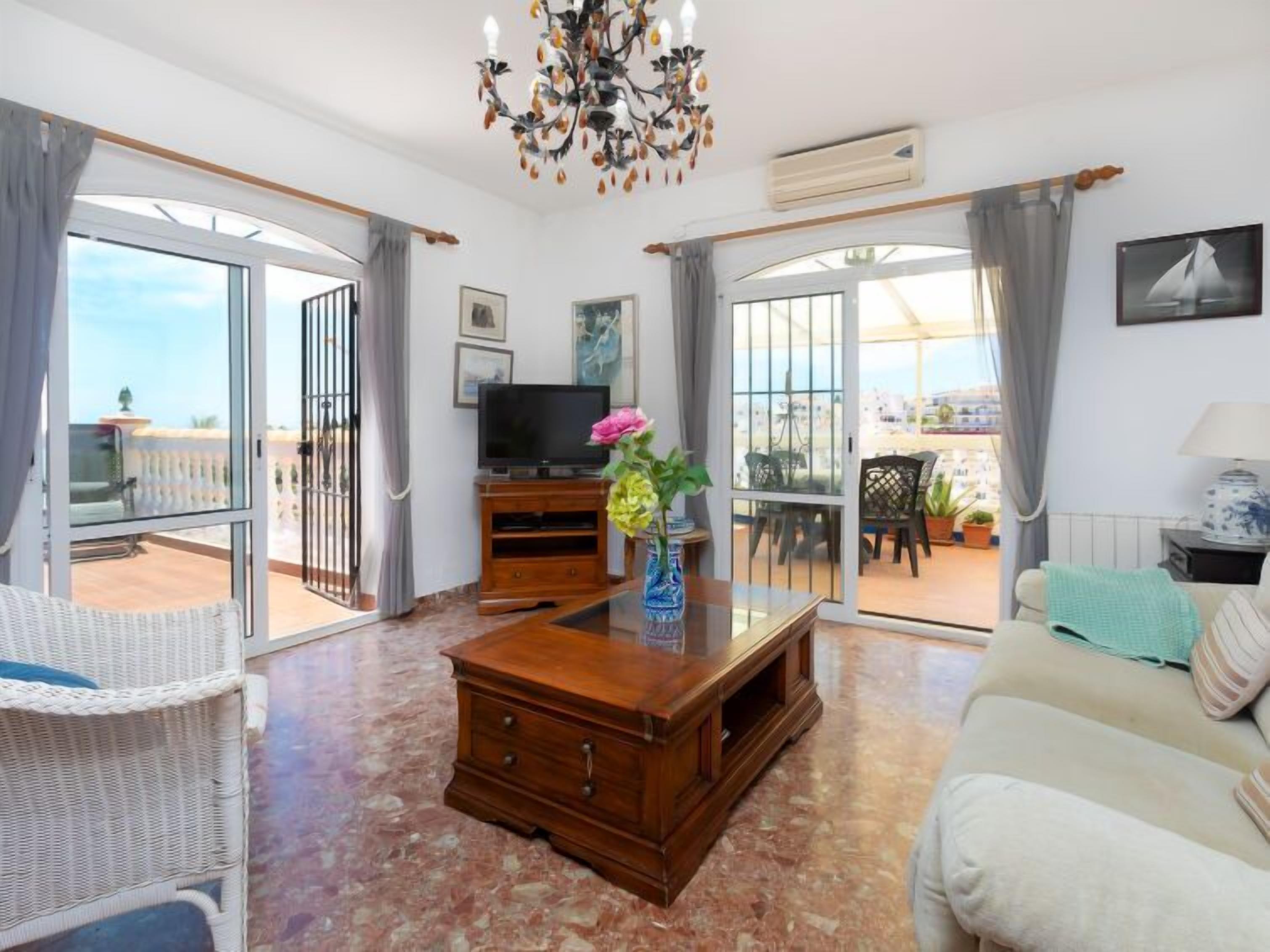 Ein geräumiges Wohnzimmer mit Fernseher, Couchtisch und Sofa. Ein Balkon bietet Blick auf das Meer.