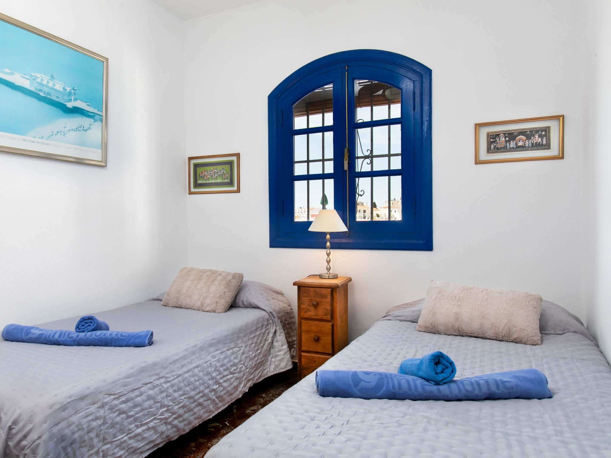 Ein Hotelzimmer mit zwei Betten, einem Holznachttisch, einer Lampe und einem großen Fenster mit blauen Rollläden.