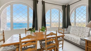Ein Zimmer mit Esstisch und Stühlen, einer Couch und großen Bogenfenstern mit Blick auf das Meer.