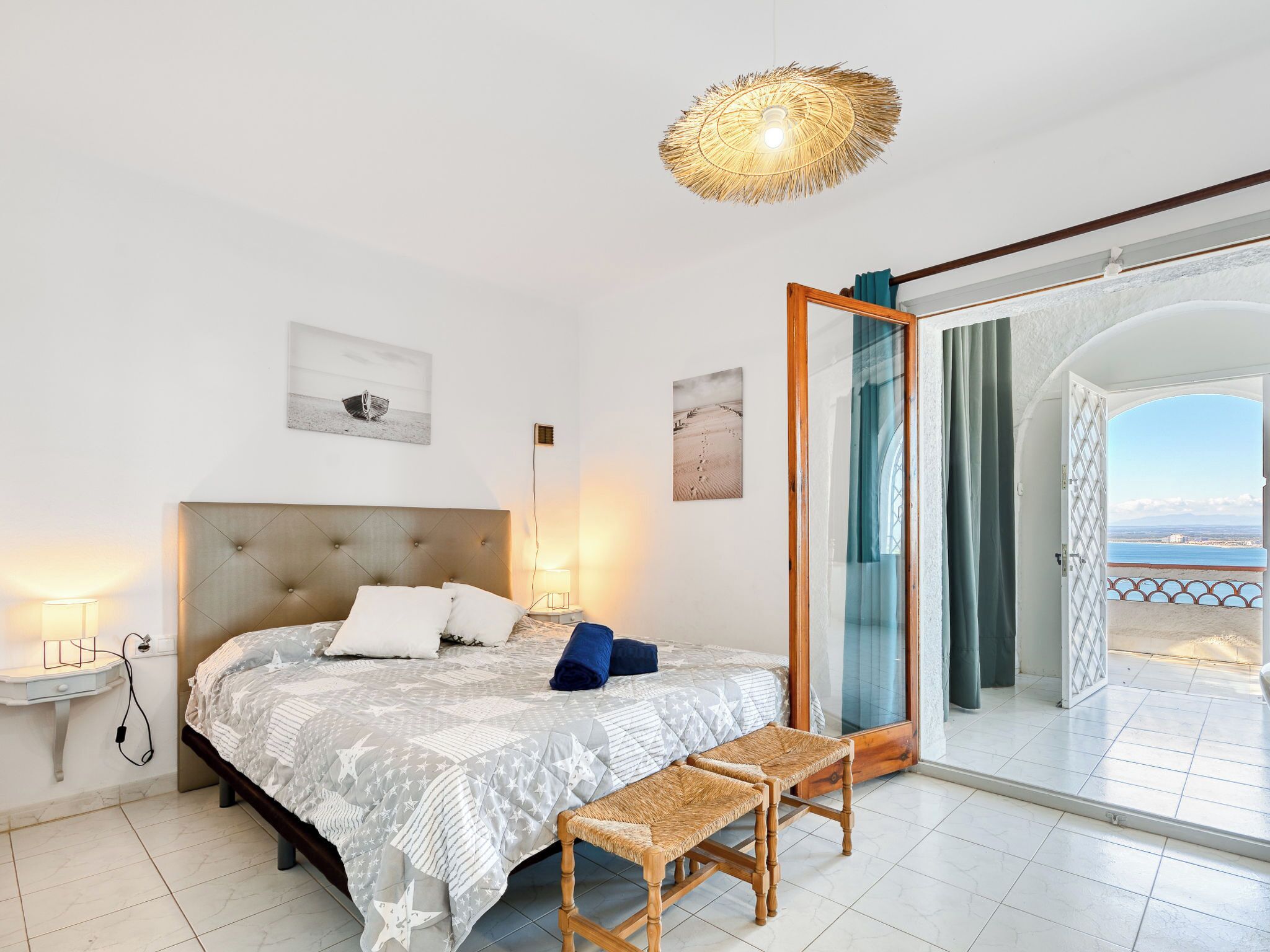 Een slaapkamer met een groot bed, een nachtkastje, een stoel, een hanglamp en uitzicht op de zee door een geopende deur.