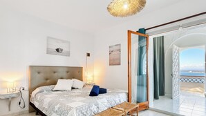 Ein Schlafzimmer mit einem großen Bett, einem Nachttisch, einem Sessel, einer Hängelampe und Meerblick durch eine offene Tür.