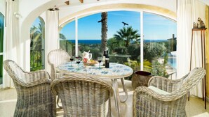 A sunlit patio with wicker furniture, a round table, and a view of the sea and palm trees. - Las Boyas by Interhome (Moraira)