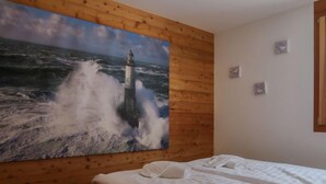 Ein Zimmer mit einem großen Gemälde eines Leuchtturms an einer Holzwand, einem Bett mit weißer Bettwäsche und einem kleinen Fenster mit Vorhängen.