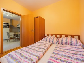Ein Schlafzimmer mit einem Bett, einem Sessel, einem kleinen Tisch und einem durch eine offene Tür einsehbarer Küchenbereich.