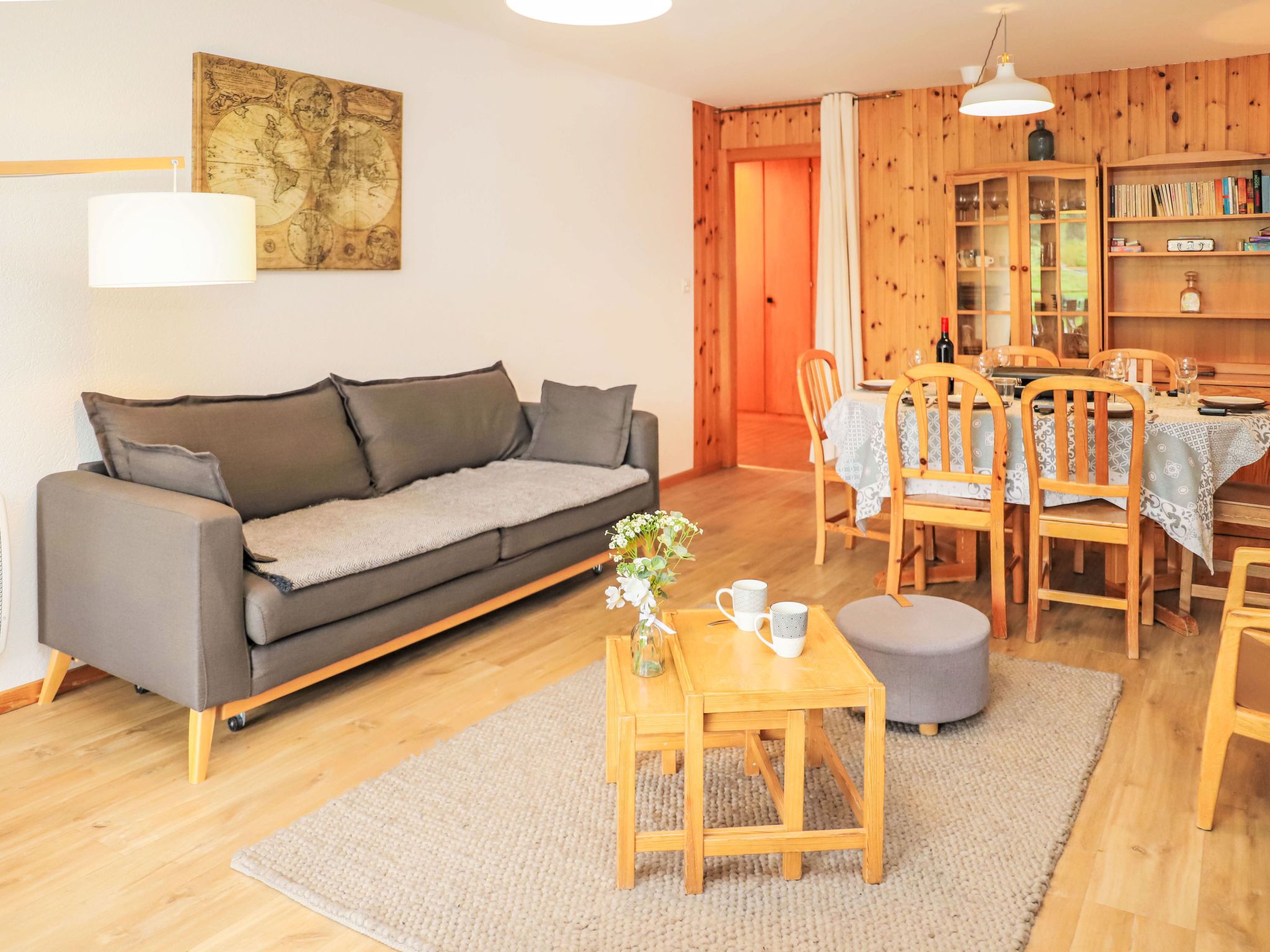 Een gezellige woonkamer met een grijze bank, een houten eettafel en stoelen. Op de muur hangt een wereldbol kunstwerk en een boekenplank vol boeken en decoratieve objecten.