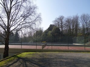 Un campo da tennis con una recinzione, alberi e un edificio sullo sfondo.