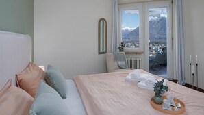 Habitación de hotel con cama, vistas a las montañas, ventana con cortinas y una pequeña mesa con toallas y un jarrón.