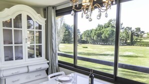 A dining area with a white table set for four, a white cabinet with glass doors, and a view of a green landscape through large windows. - Les Catalanes du Golf by Interhome (Saint Cyprien)