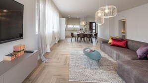 Ein modernes Wohnzimmer mit Flachbildfernseher, einer Couch, einem Couchtisch und einem Essbereich im Hintergrund.