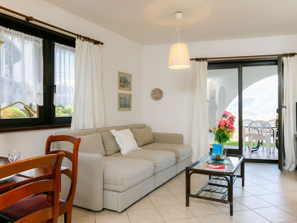 A cozy living room with a sofa, a glass coffee table, and a dining area with chairs. There is a window with curtains, a ceiling light, and a view of the outdoors.