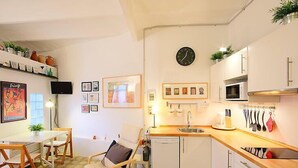 Een compacte keuken met witte kasten, een magnetron, een spoelbak en een klok aan de muur. Er staan fotolijstjes en een klein plantje op de planken.
