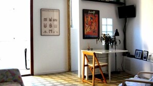 Een kleine, goed verlichte kamer met een wit bureau, een stoel, een bed en een televisie op een standaard. Er hangen afbeeldingen in lijsten aan de muur en staat een plant in een pot op het bureau.