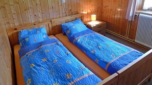 A wooden-paneled room with two beds, each with blue floral bedding. There is a nightstand with a lamp and a framed picture on the wall.