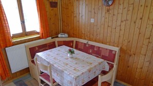 A wooden-paneled room with a table covered in a floral tablecloth, two chairs, a clock on the wall, and a window with curtains.