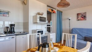 Eine kompakte Küche mit Kaffeemaschine, Mikrowelle und kleinem Kühlschrank. Im Wohnbereich befindet sich ein Esstisch mit Stühlen sowie ein blaues Sofa.