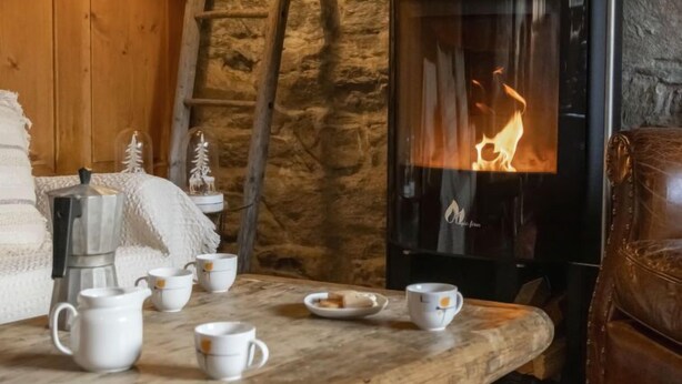 A cozy room with a stone wall, a wooden table with cups and a kettle, and a fireplace with a visible fire. - La Pive by Interhome (Verbier)