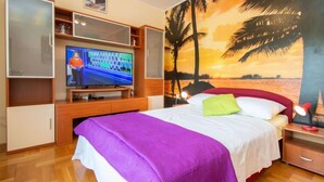 Quarto de hotel com cama, televisão, móveis de madeira e um mural com pôr do sol na parede.