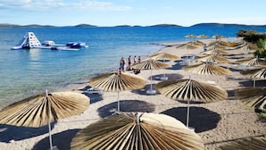 Uma praia com guarda-sóis e espreguiçadeiras, um barco e pessoas caminhando pela orla.