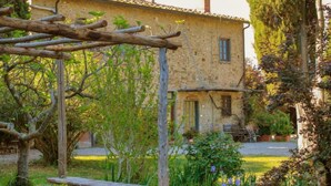 Un bâtiment en pierre avec un toit en tuiles, une pergola en bois et un jardin où poussent différentes plantes et fleurs.