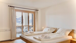 A double bed with white linens and towels, a wooden headboard, a bedside lamp, and a view of a snowy landscape through a large window with curtains.