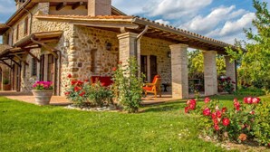 Une maison en pierre dotée d’un patio couvert, d’une cheminée et d’un jardin agrémenté de fleurs rouges.