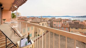 Ein Balkon mit Tisch für zwei Personen, mit Blick auf eine Küstenstadt mit terrakottafarbenen Gebäuden und Meerblick.