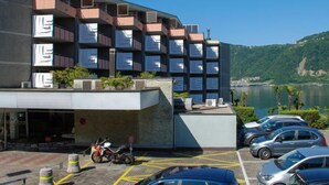 A modern building with multiple balconies, a parking area with cars, and a view of a lake and hills. - App. 511 by Interhome (Bissone)